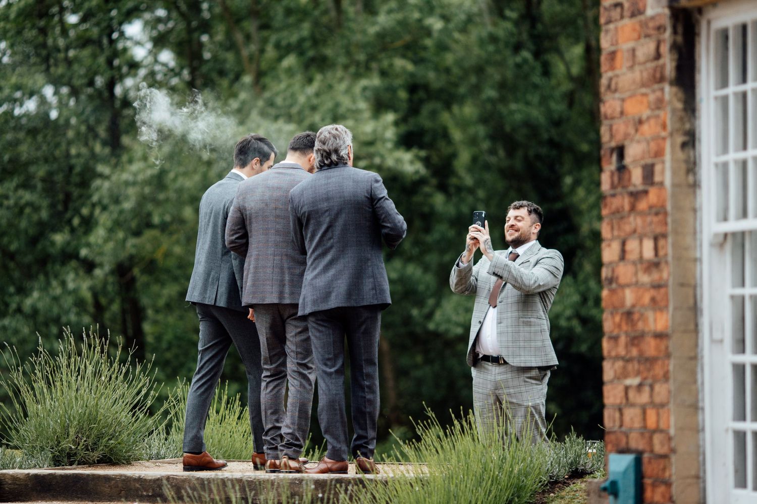 Wedding party group gathered on lawn outside brick building with white windows.