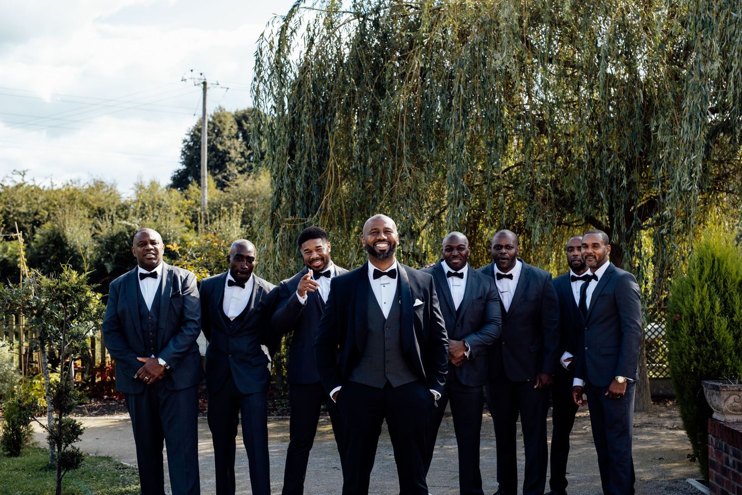 Groomsmen in black tuxedos line up for formal photo under willow trees.