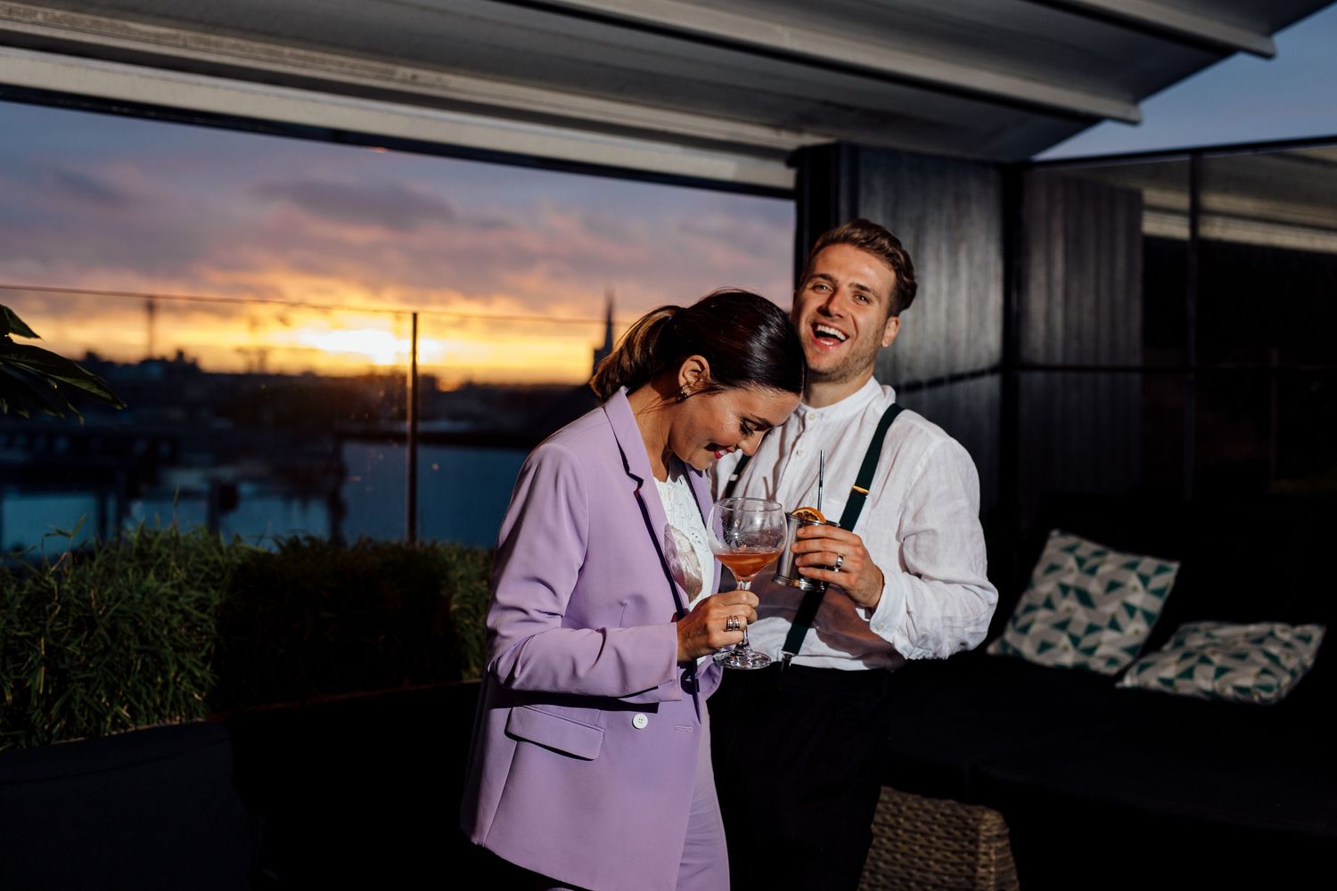 A couple shares drinks on a balcony during a vibrant sunset.
