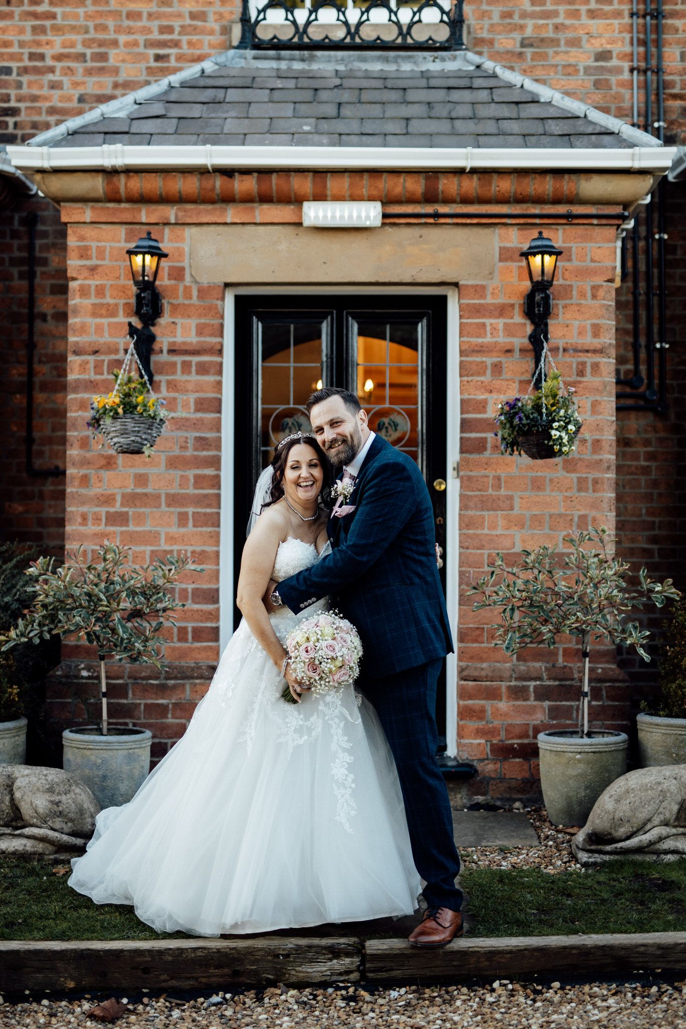 A newlywed couple poses in front of a brick entrance with hanging lanterns and potted plants.