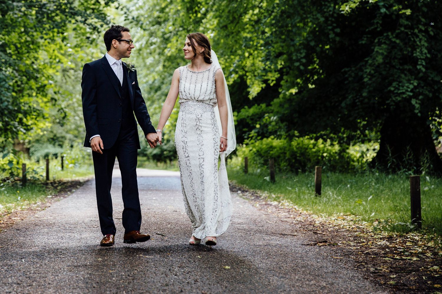 Newlyweds walking hand in hand down a peaceful garden pathway surrounded by trees.