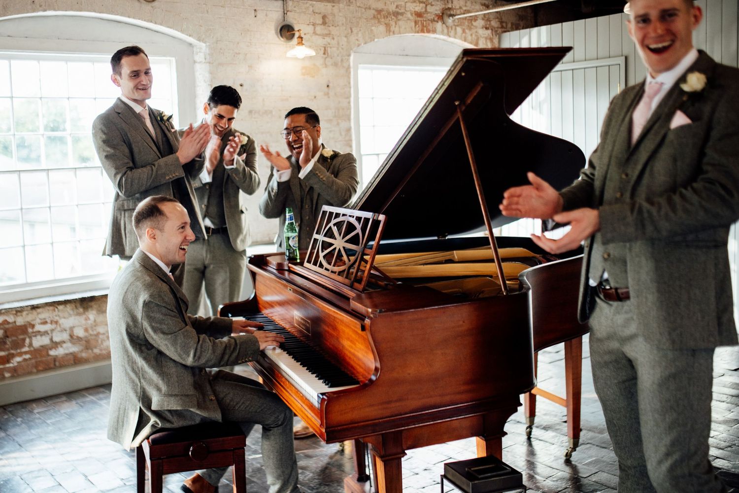 Groomsmen gather around vintage piano in rustic brick-walled venue.