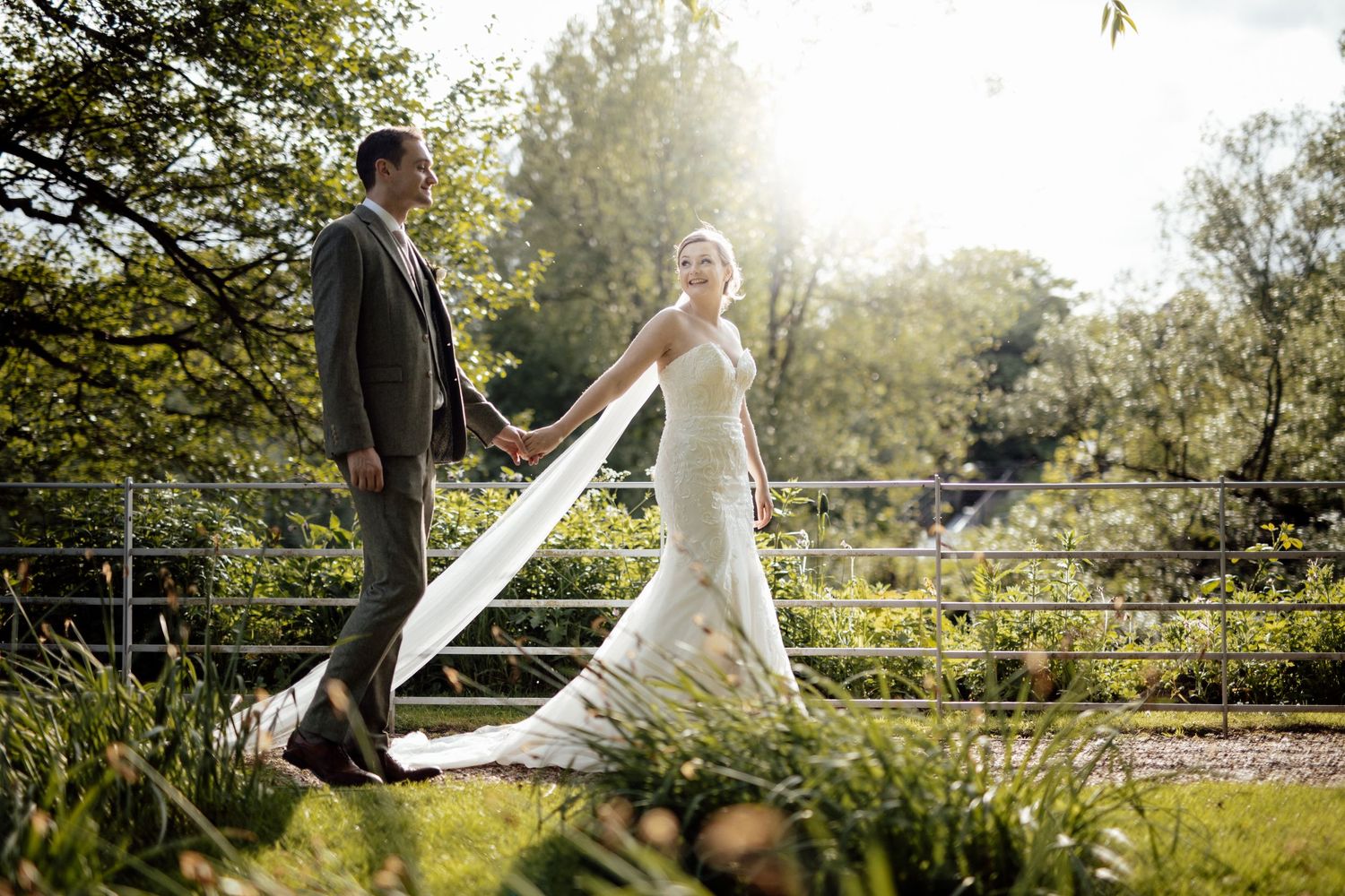 A wedding couple poses together in a sunlit outdoor garden setting with lush greenery.
