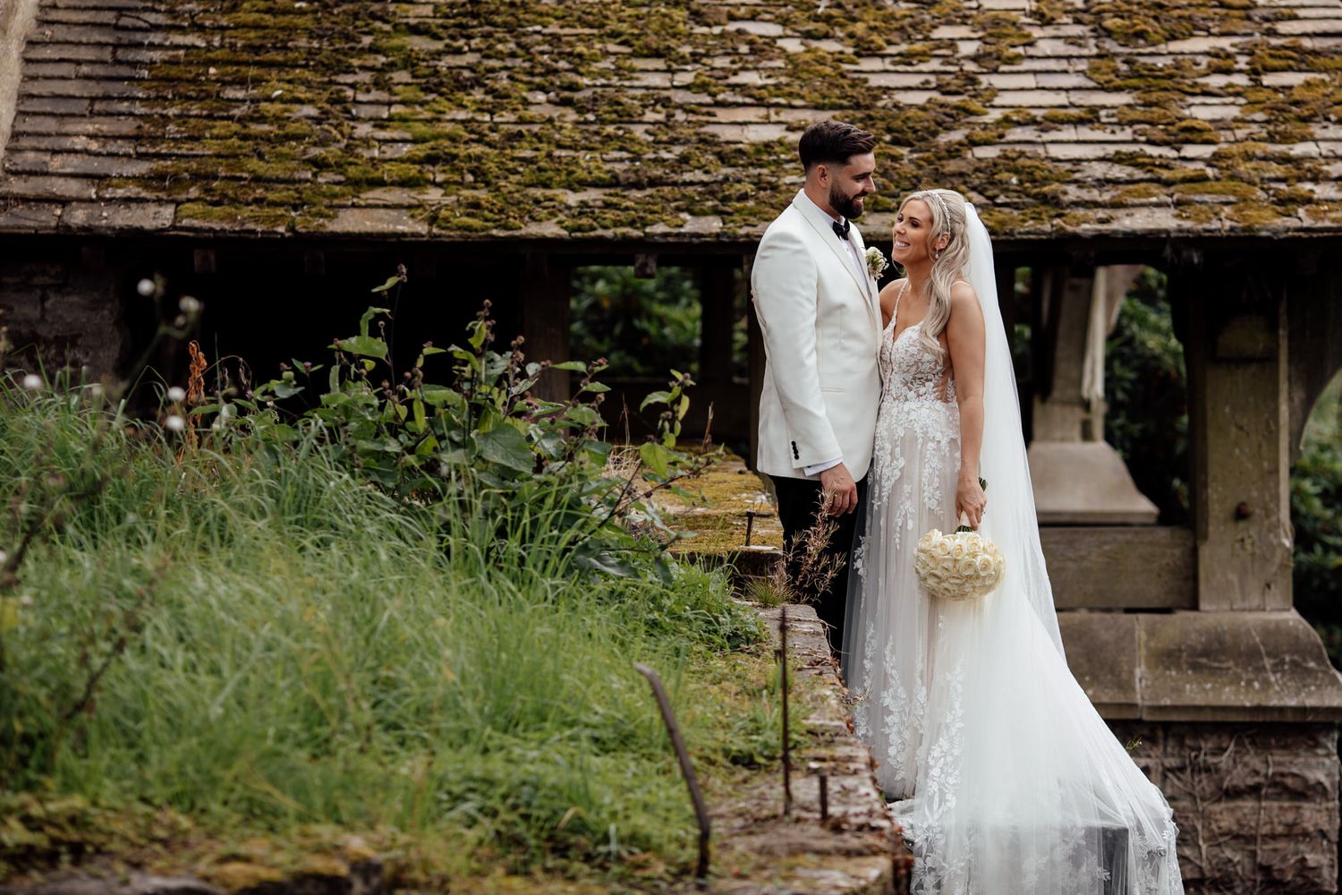 Bride in flowing white dress walks through rustic stone archway.