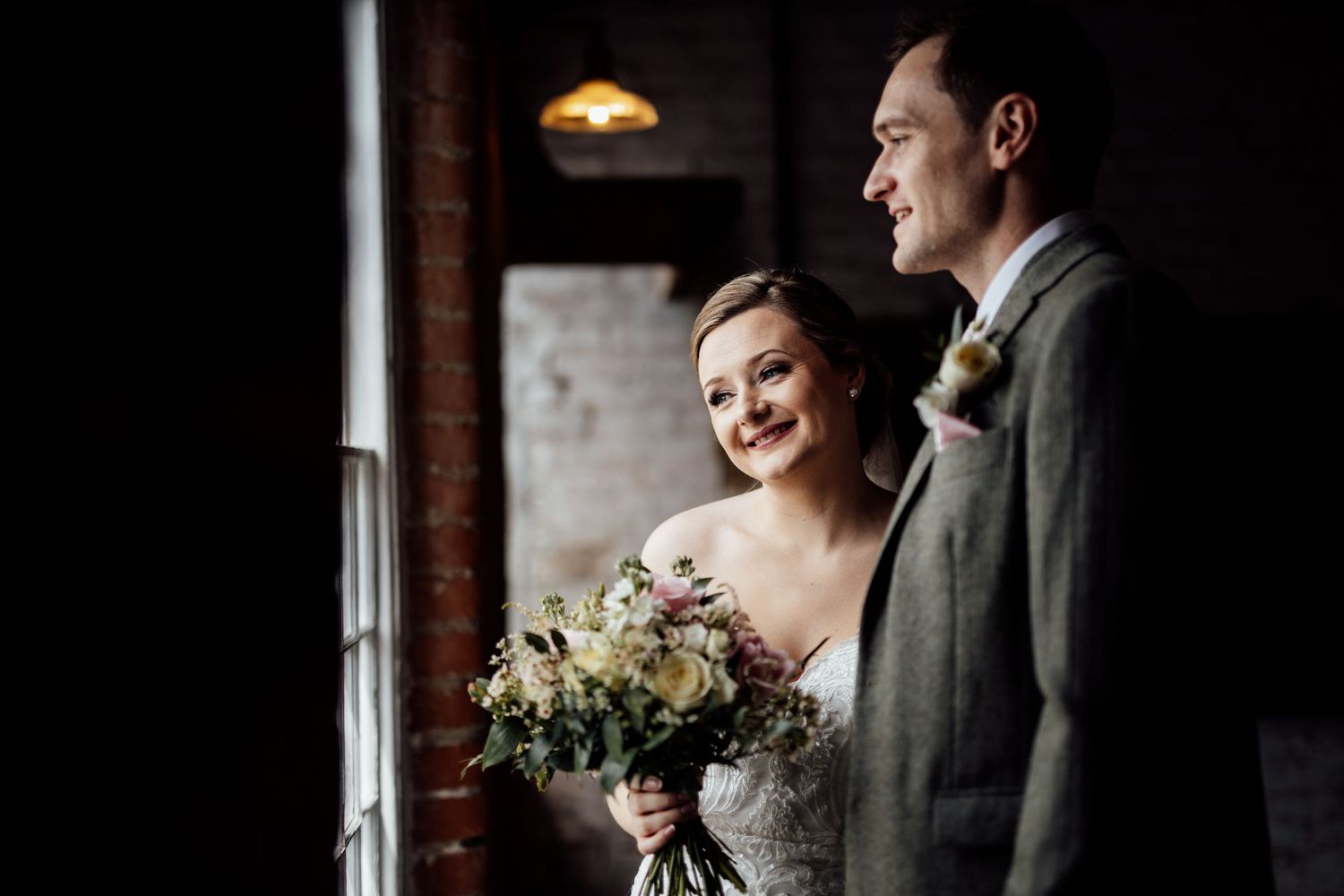 Newlyweds share intimate moment by window in industrial brick venue.