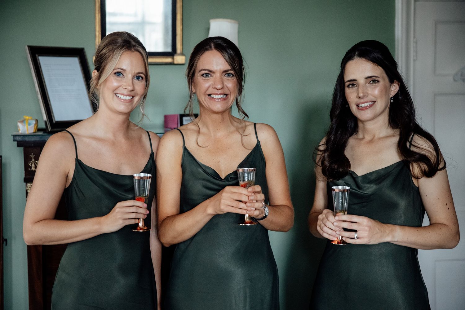 Bridesmaids in black dresses toasting with champagne glasses against a mint green wall.