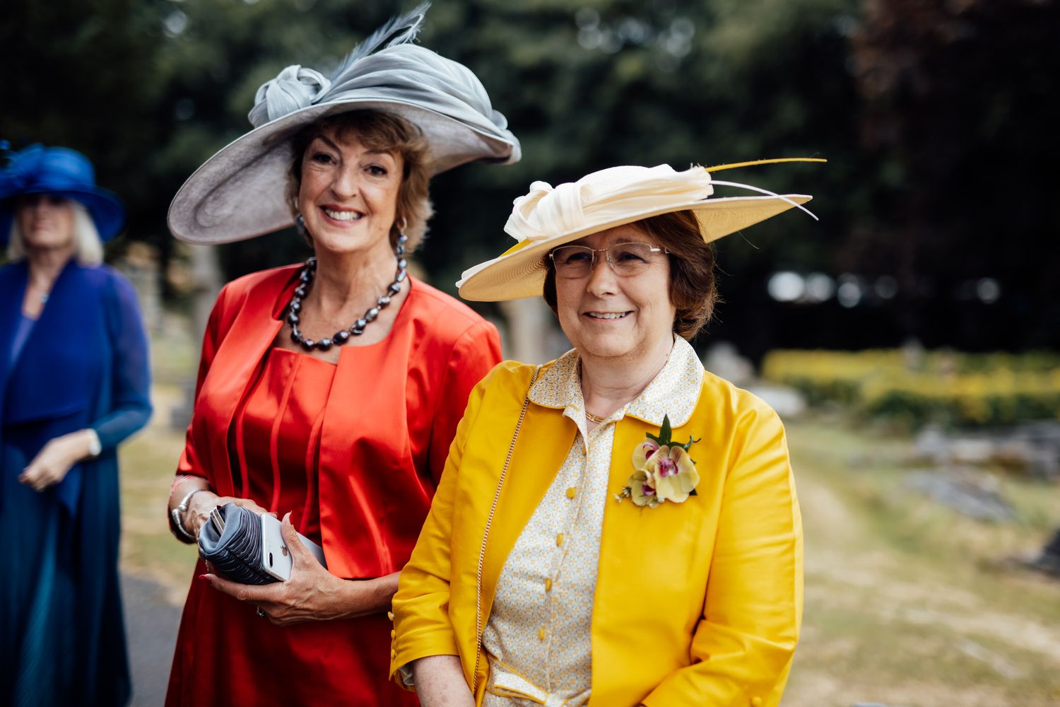 Wedding guests in bright formal attire and fancy hats enjoying an outdoor celebration.