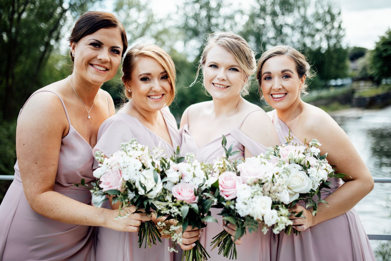 Bridesmaids in blush dresses holding white floral bouquets pose together outdoors.