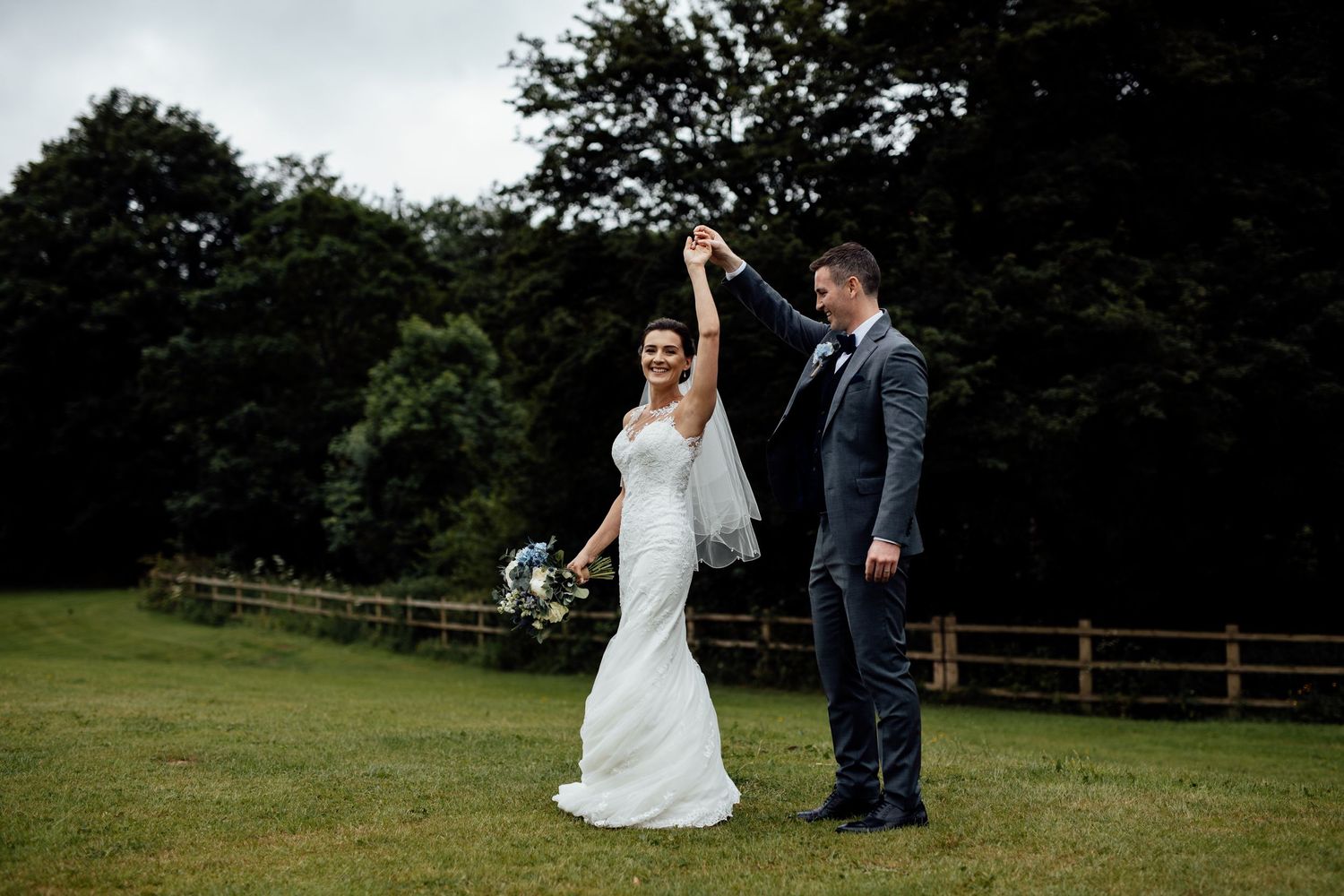 Wedding couple walks hand in hand across a scenic field with trees in the background.