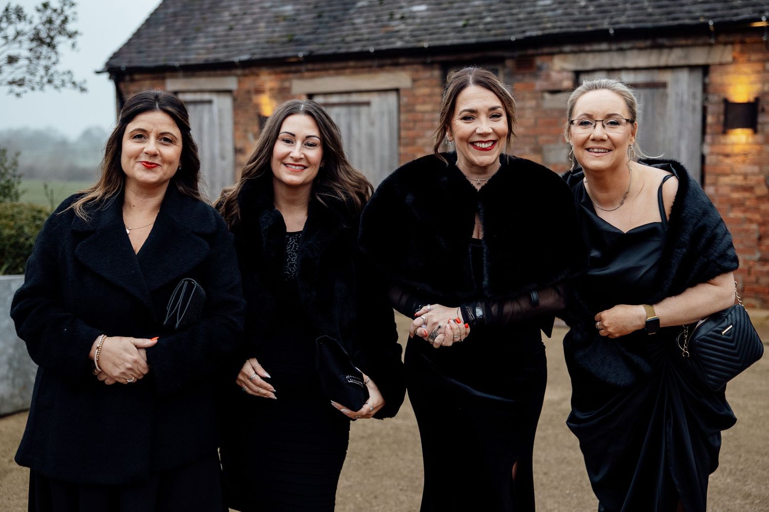 Wedding party members in black dresses pose together against a rustic backdrop.
