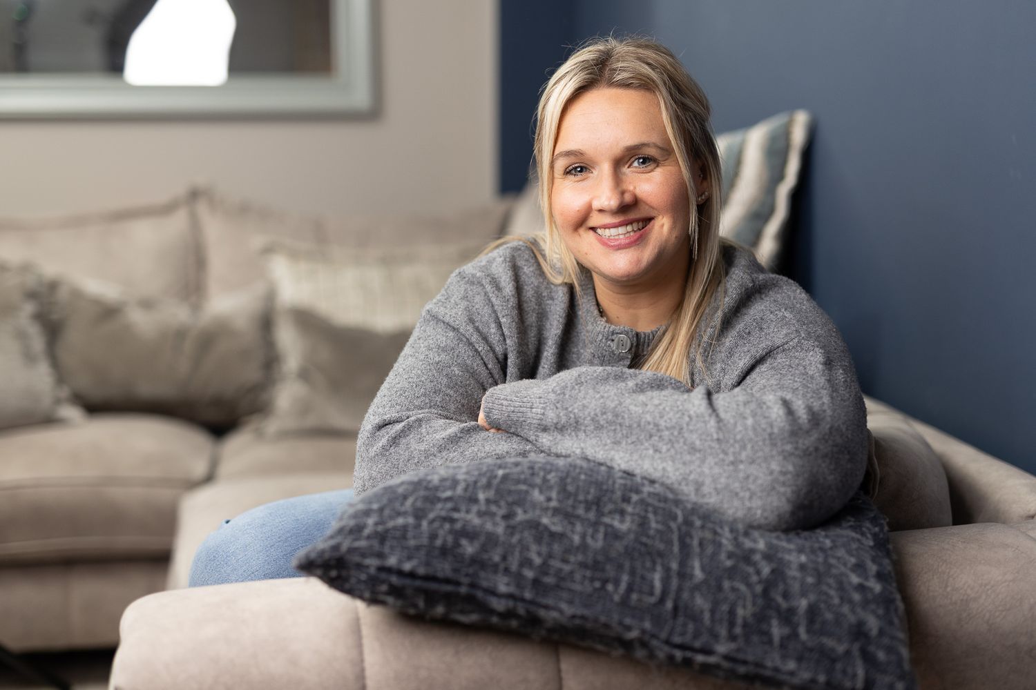Smiling blonde woman relaxing on a beige sofa, hugging a grey pillow in a cozy living room.