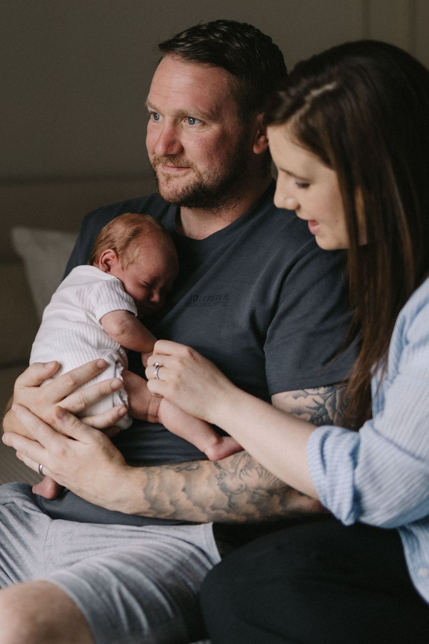 A tattooed father holds a sleeping newborn baby while the mother looks on tenderly.