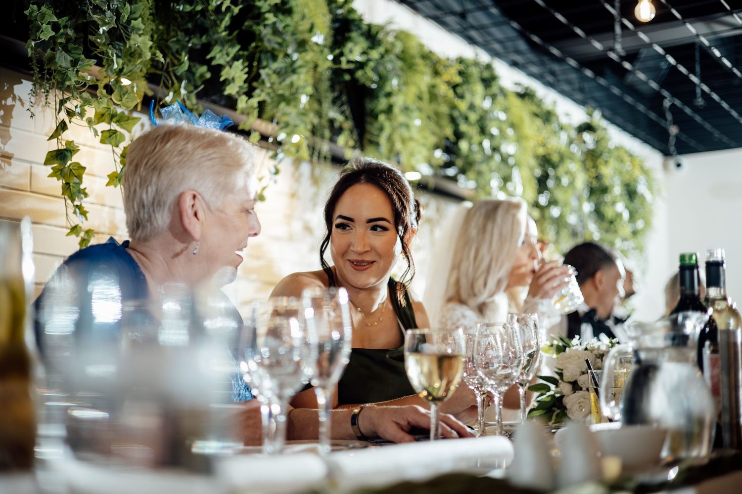 Women socializing at an elegant dinner party with wine glasses and lush green ivy decor.