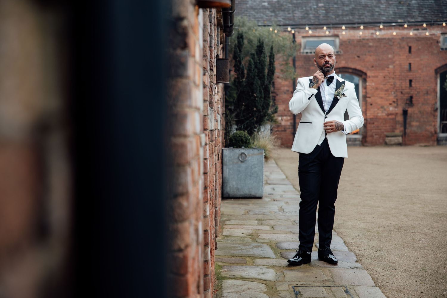 A person in a white tuxedo jacket stands against a brick wall outdoors.