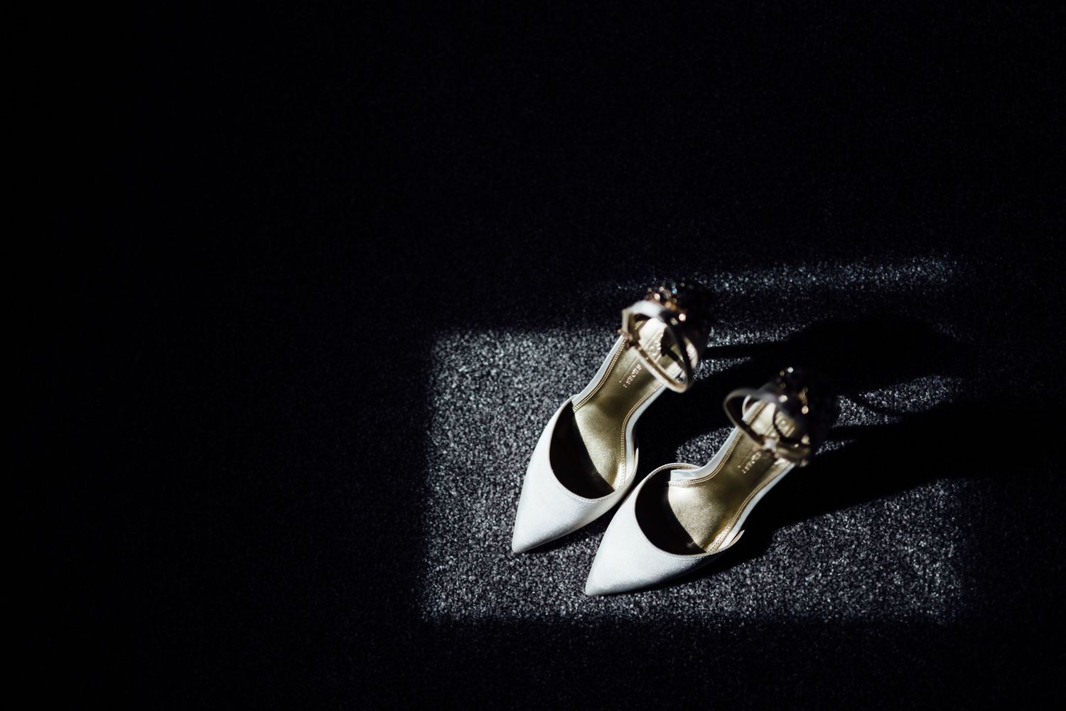 Elegant white bridal shoes captured in dramatic lighting on dark background.