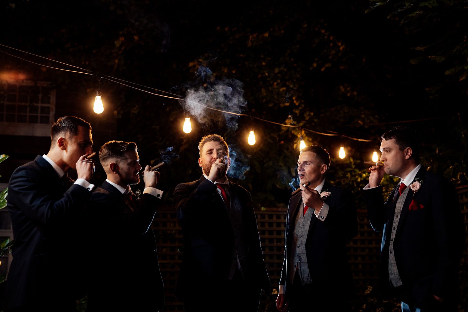 Wedding reception guests mingling under string lights in dark atmospheric setting.