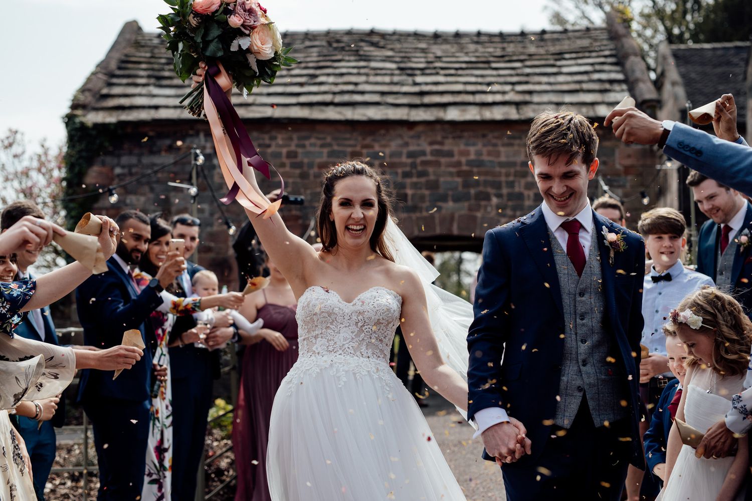 Wedding guests throw confetti over celebrating newlyweds outside brick building.