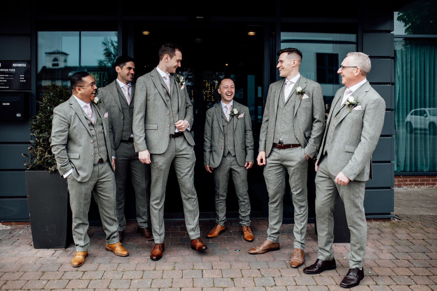Groomsmen in matching light gray suits and brown shoes stand casually together outside a building.