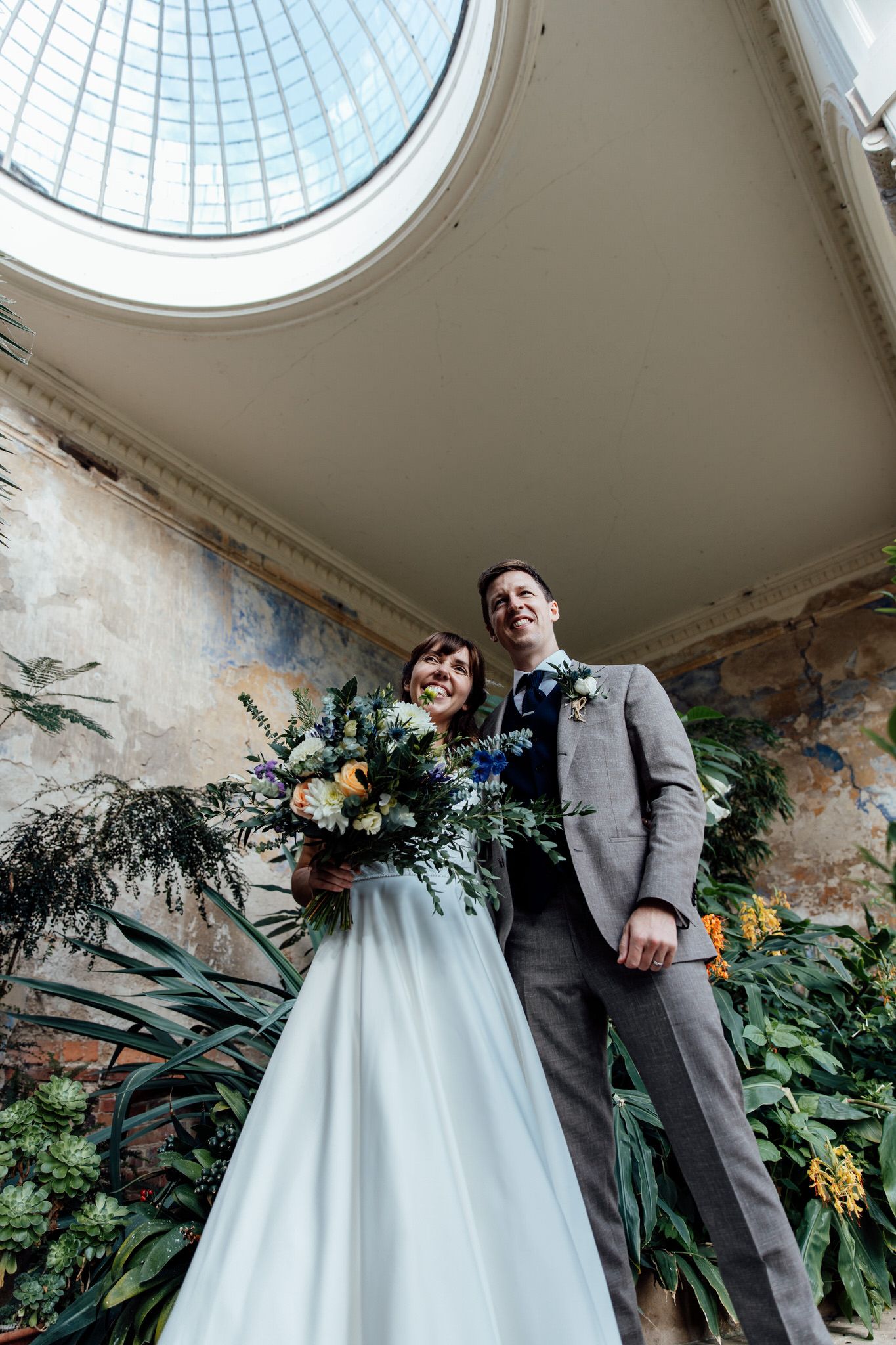 Wedding couple standing beneath glass dome ceiling surrounded by lush greenery and flowers.