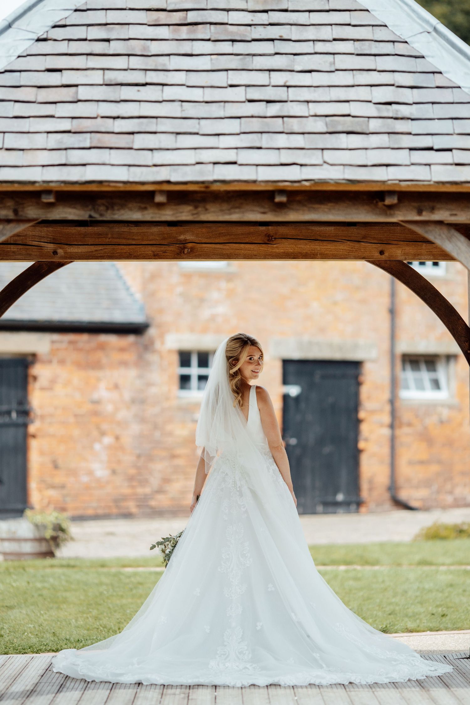 Flowing white wedding dress displayed against rustic brick building and wooden gazebo.