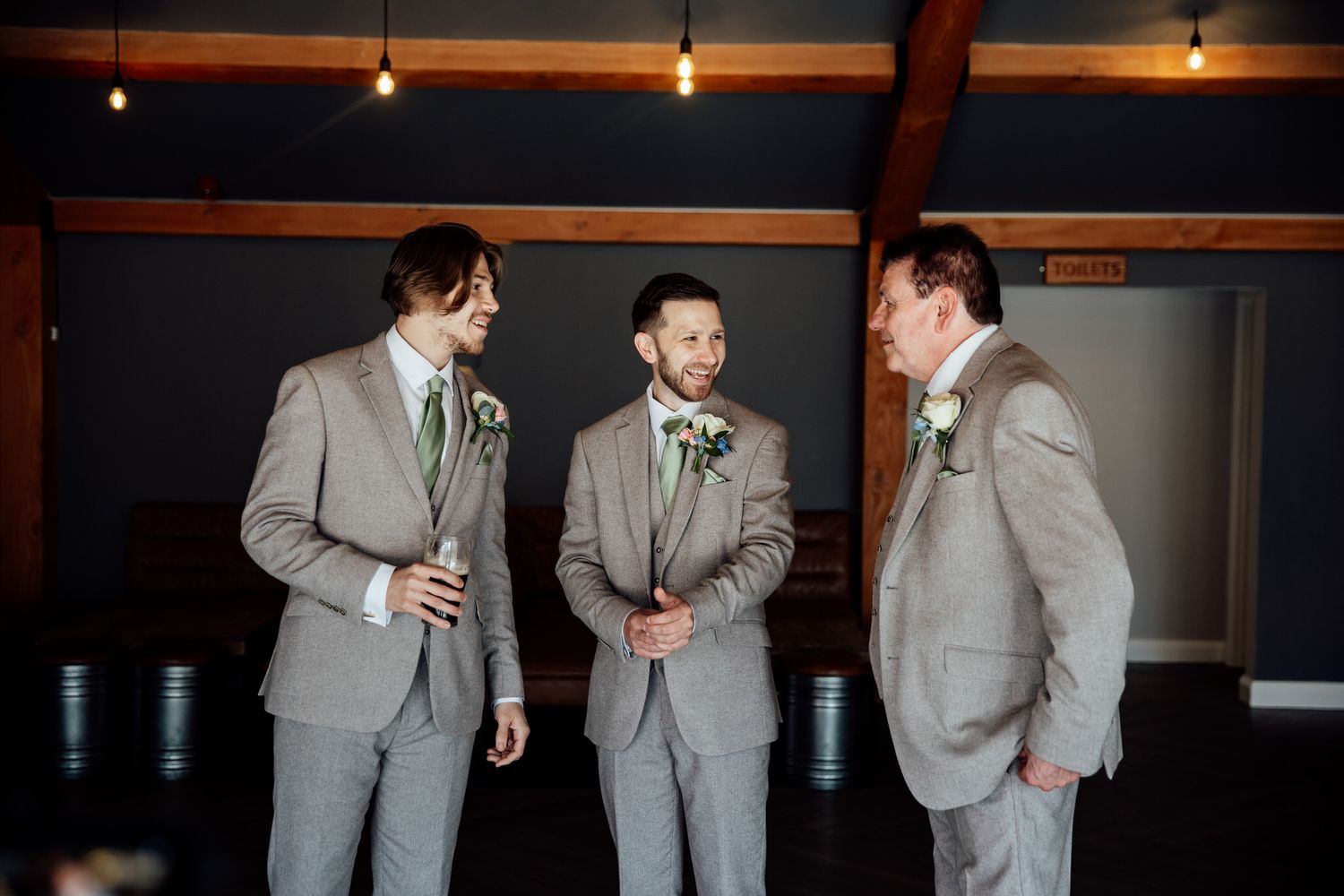 Groomsmen in light gray suits laughing and joking together at an indoor venue.
