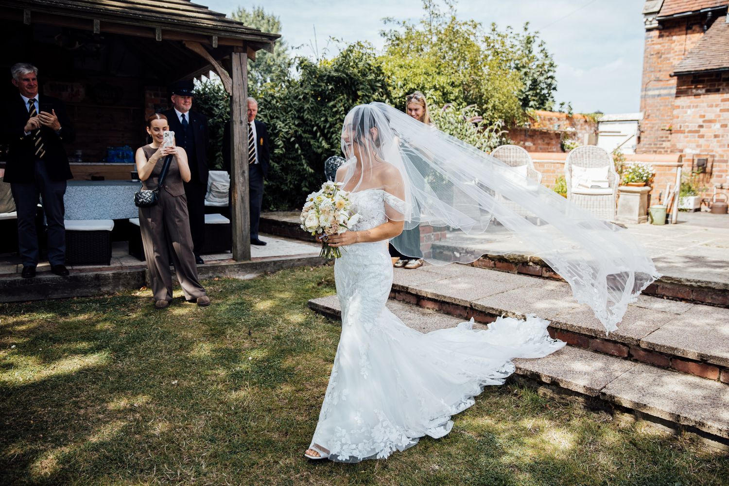 Bride's flowing white dress and veil trail behind her in an outdoor garden setting.