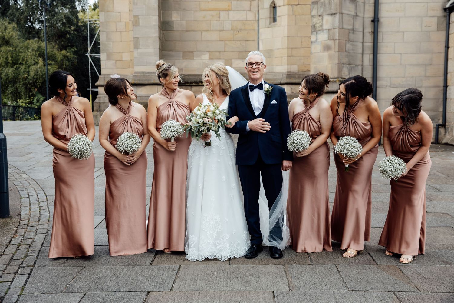 Wedding party poses in formal attire outside a stone building with pink bridesmaid dresses.