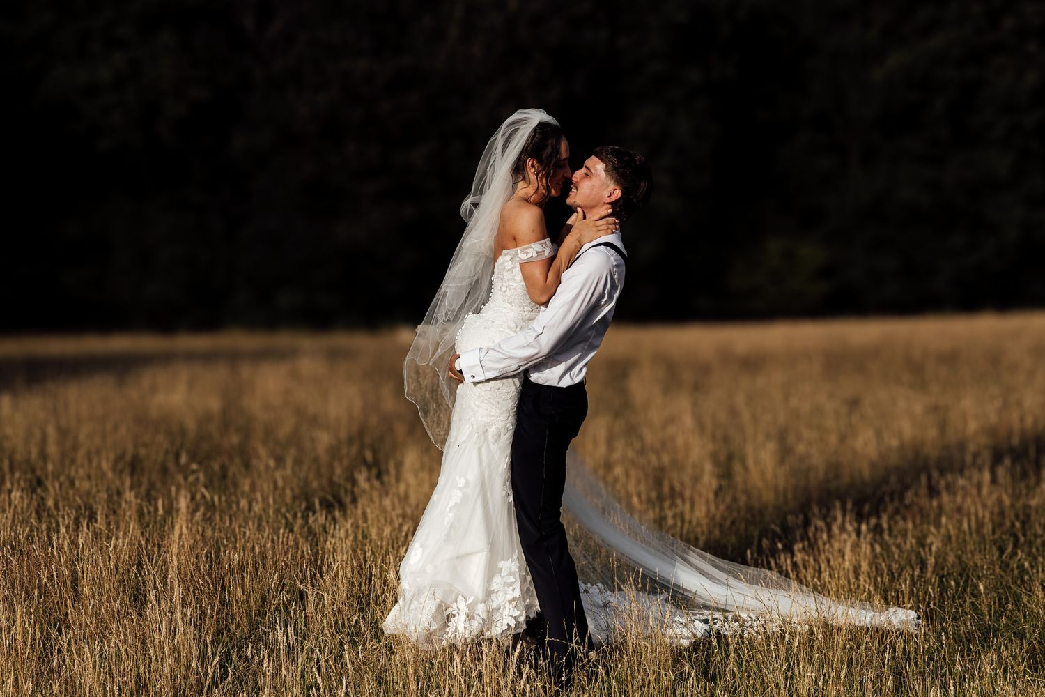 Romantic wedding couple embraces in a golden field at sunset with flowing veil and dress.