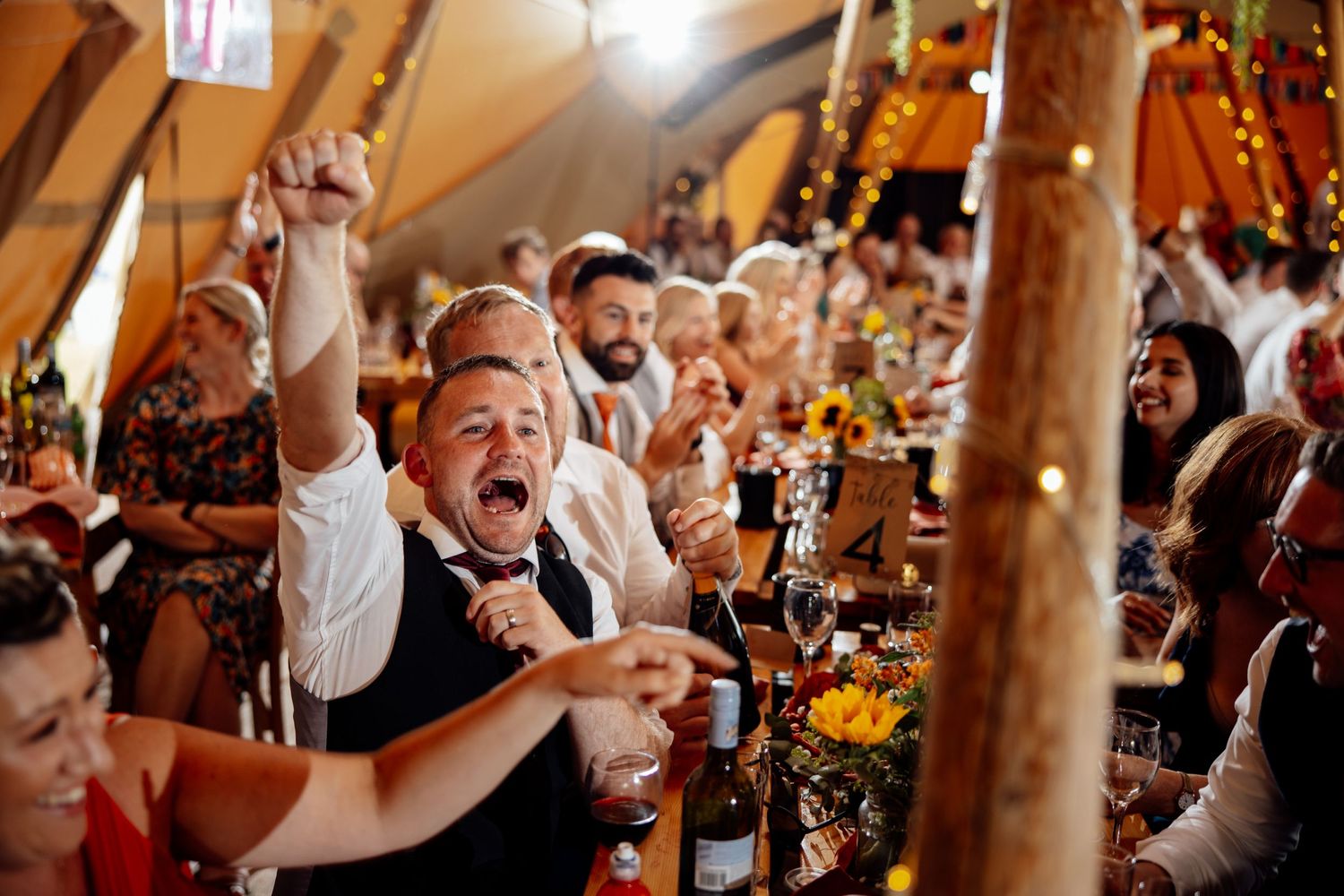 Wedding reception guests raise their glasses in celebration inside a rustic wooden venue.