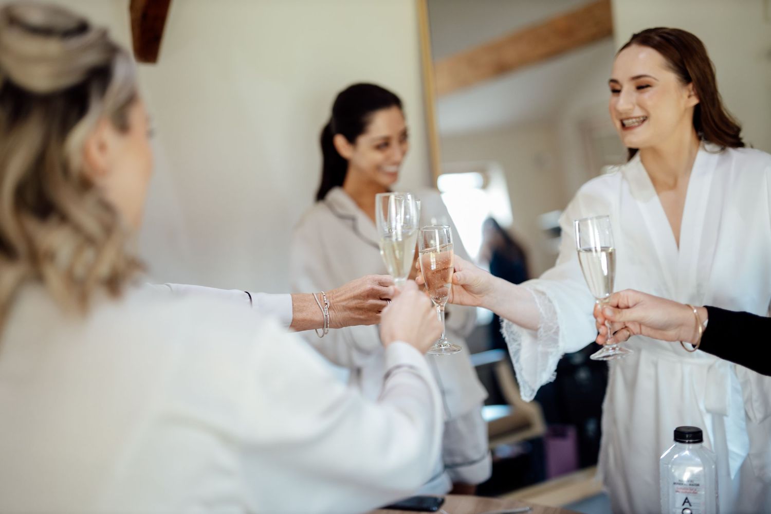 Women in white robes toasting with champagne glasses during wedding preparations.
