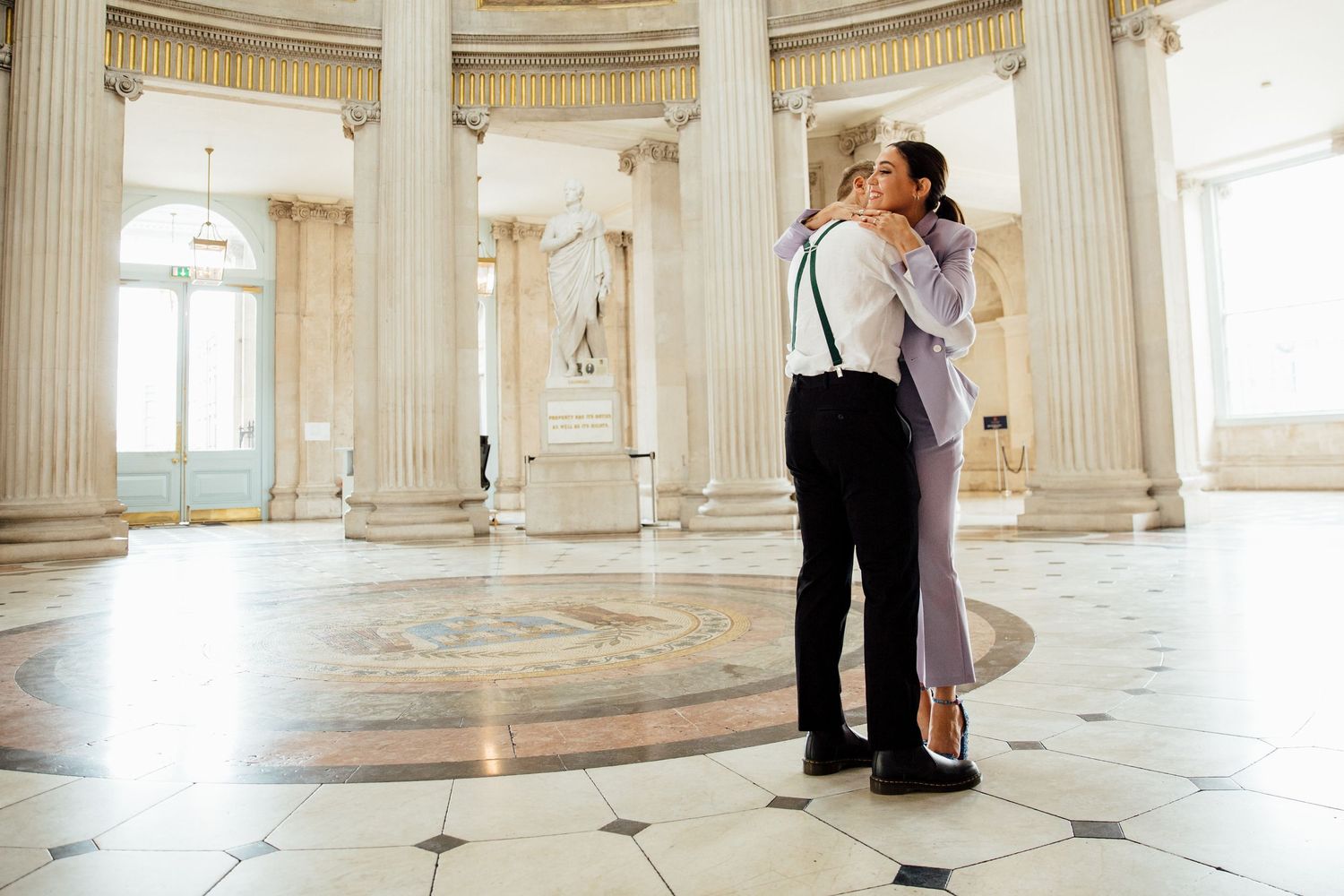 Elegant couple embracing in grand white marble hall with classical columns and natural light.