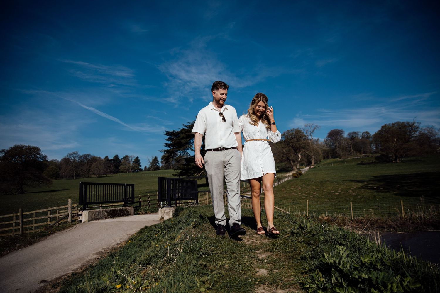 A couple in white outfits walking together on a scenic country path during a sunny day.