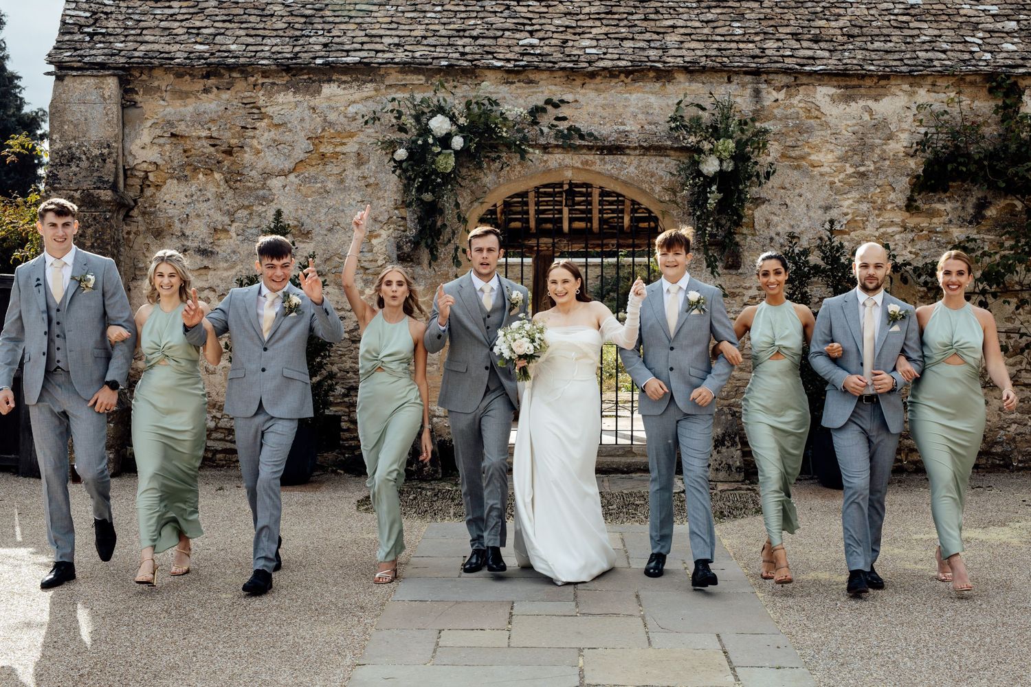 Wedding party sitting together on stone steps of rustic venue wearing grey and sage green attire.