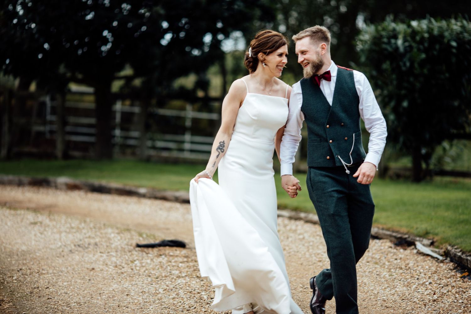 Newlyweds walk together and laugh on a tree-lined path in formal attire.