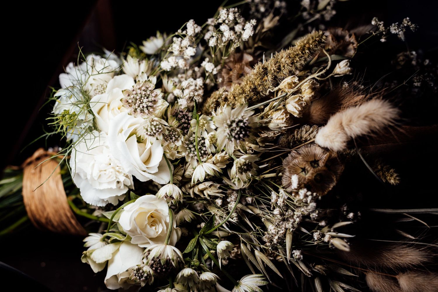 Elegant white and cream floral wedding bouquet with roses and dried elements on dark background.