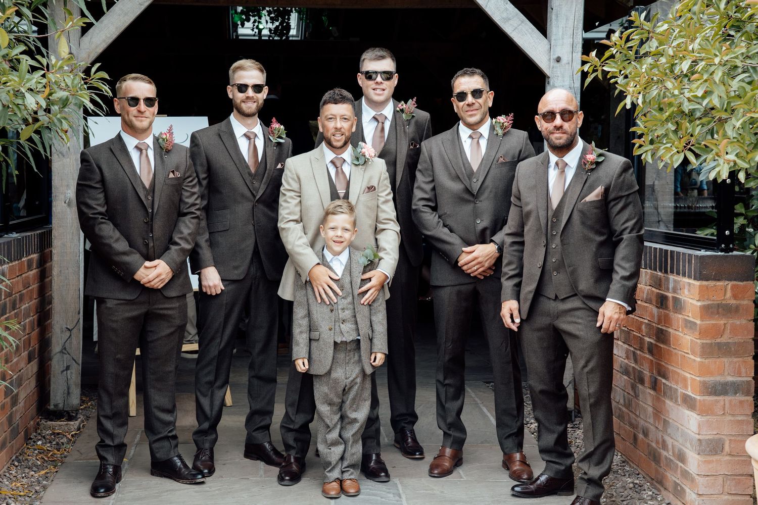 Groomsmen in matching gray suits and sunglasses pose with a young boy in a suit at an outdoor wedding venue.
