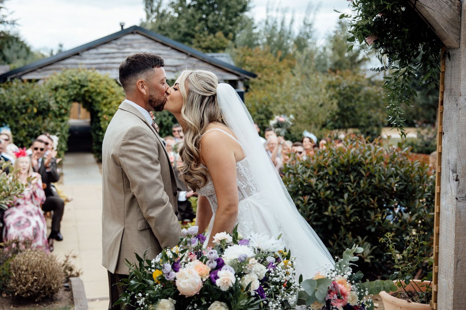 Bride and groom share first kiss at outdoor garden wedding ceremony surrounded by guests and colorful floral bouquet.