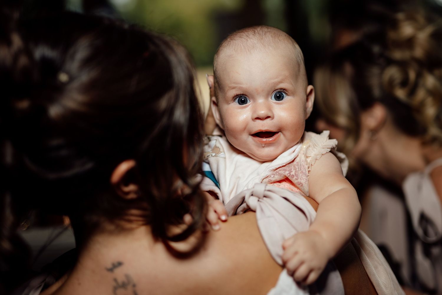 Adorable smiling baby with blue eyes being held up at an outdoor wedding.
