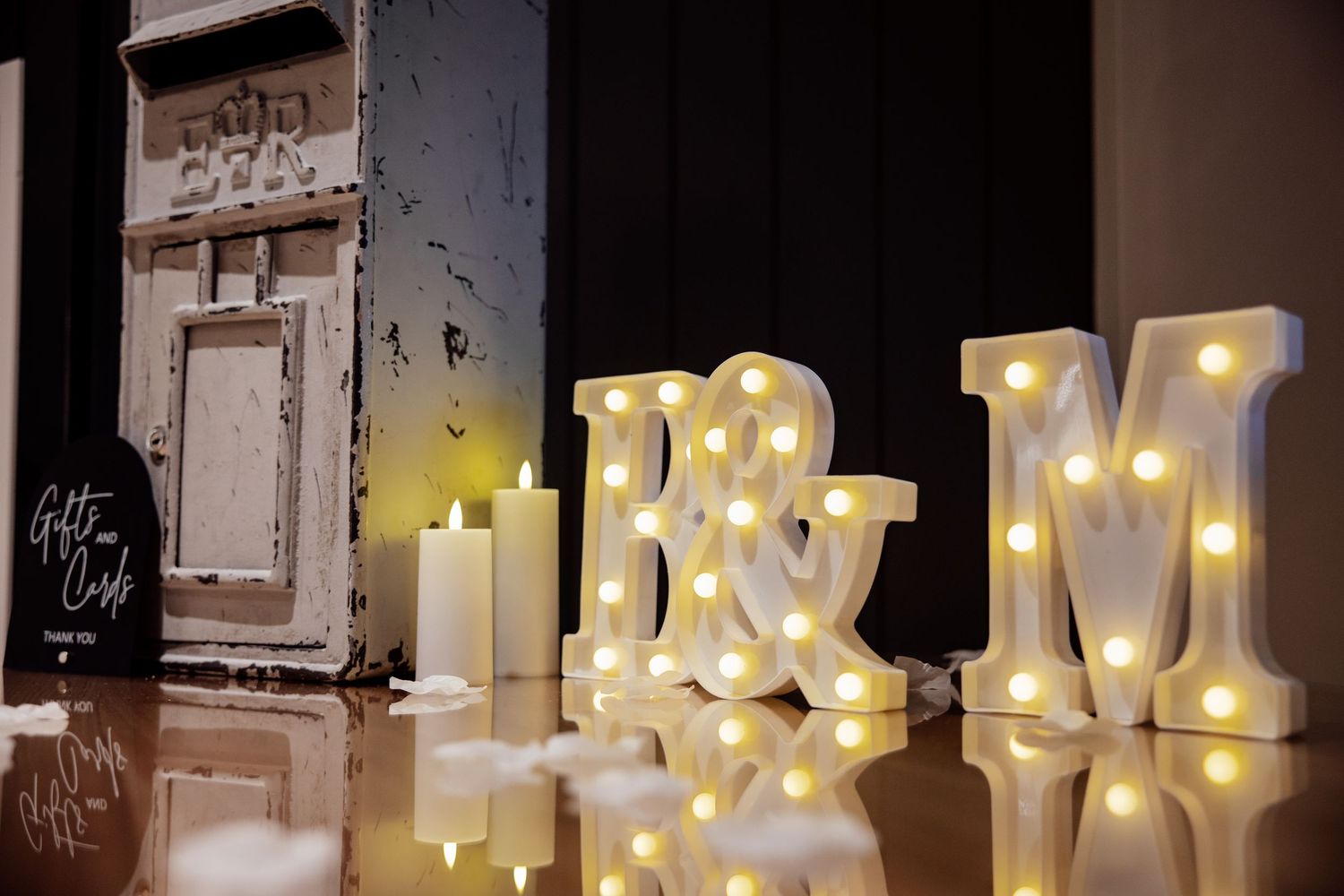 Illuminated marquee letters B&M with LED lights beside white candles on a reflective surface.