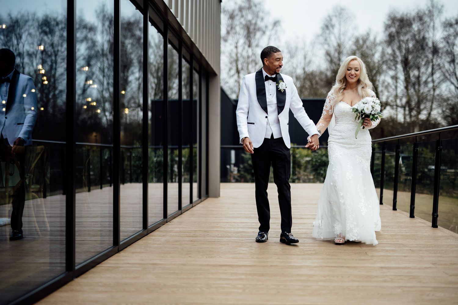Bride and groom holding hands and walking on a wooden deck outside a modern glass wedding venue.