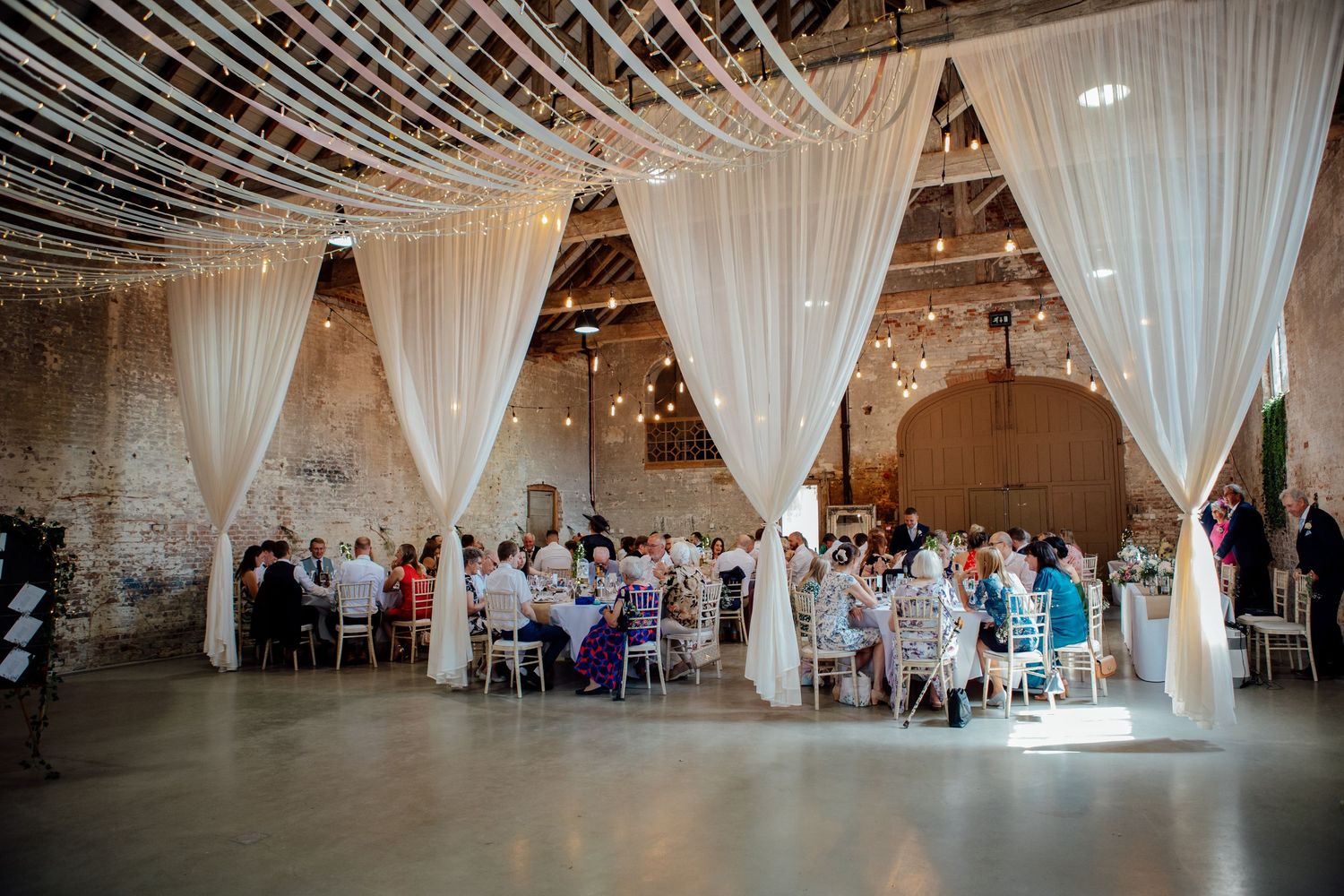 Panoramic view of an elegant wedding reception venue with white drapes and twinkling string lights overhead.