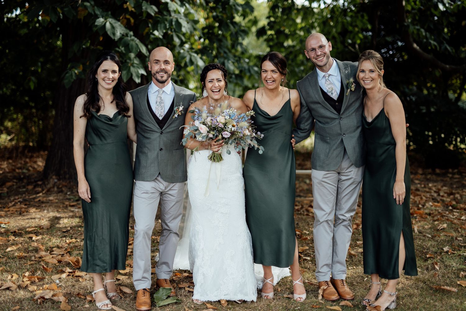 Wedding party in coordinated gray and dark green attire posing together outdoors.