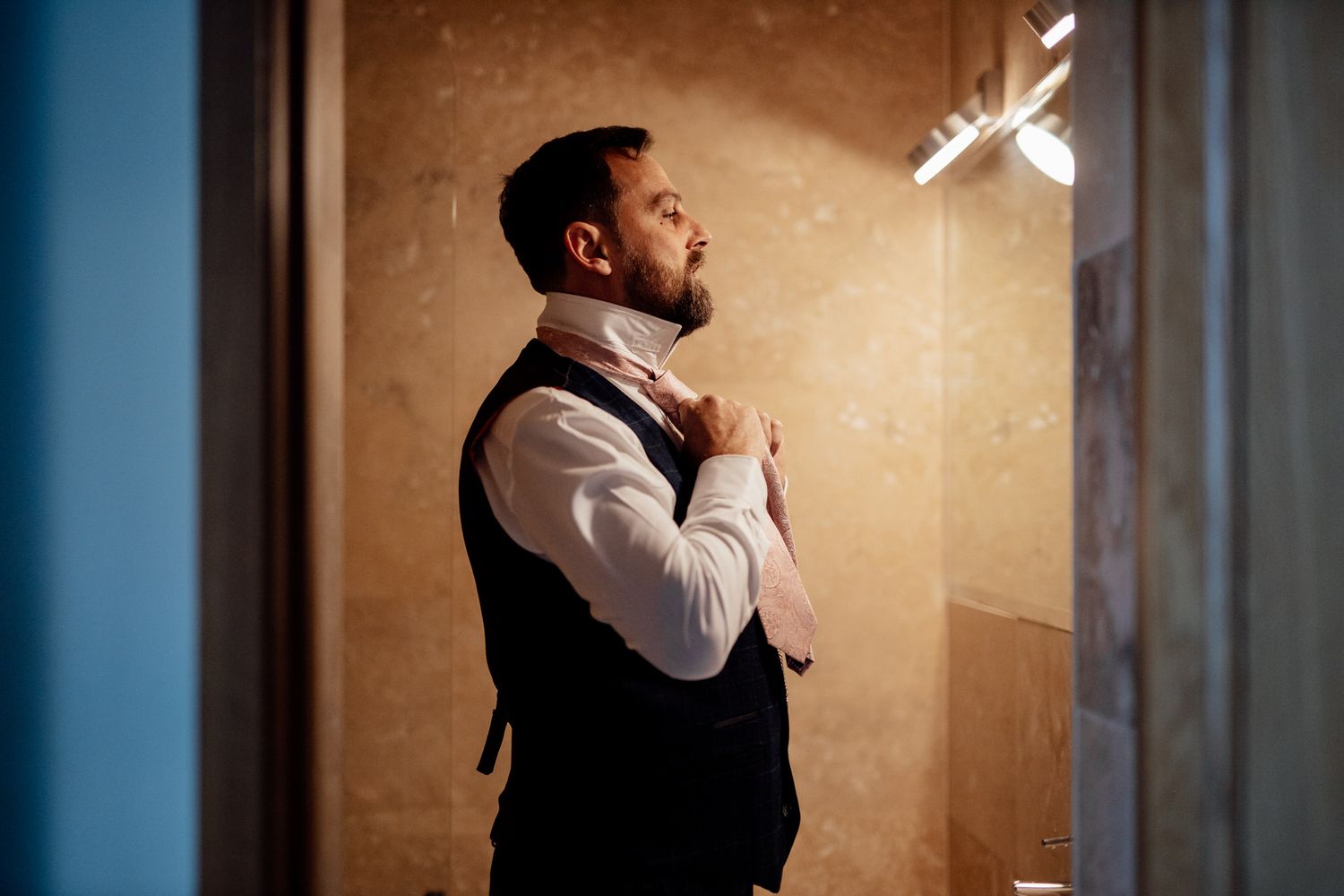 Dramatic side-lit portrait of groom getting ready in formal attire against warm backdrop.
