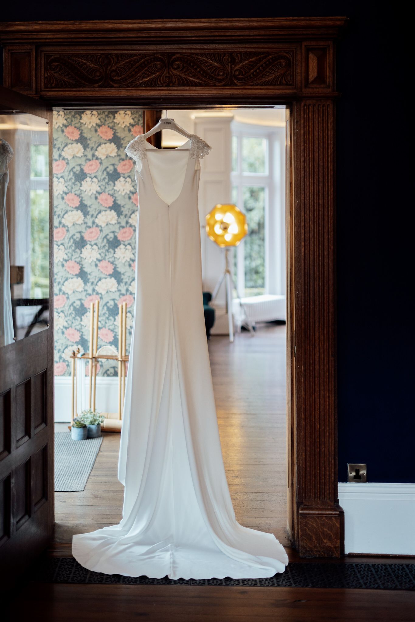 A white wedding dress hanging in a room with floral wallpaper and natural lighting.