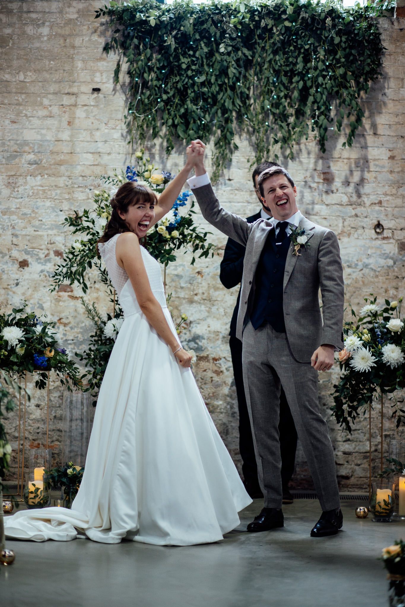 Wedding couple celebrating with joyful movements against a decorated brick wall with greenery.