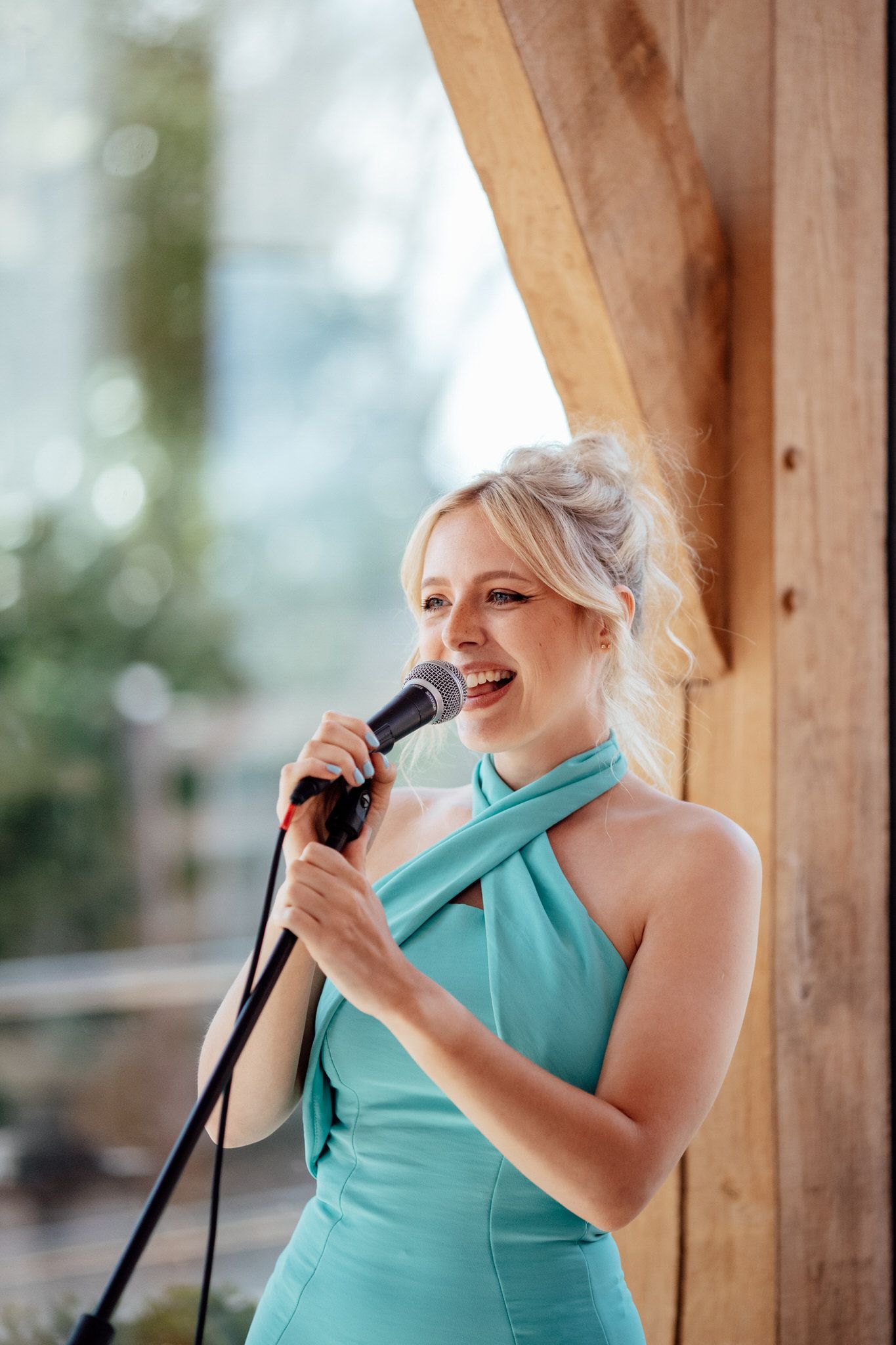 Person in turquoise dress singing with microphone in wooden pavilion setting.