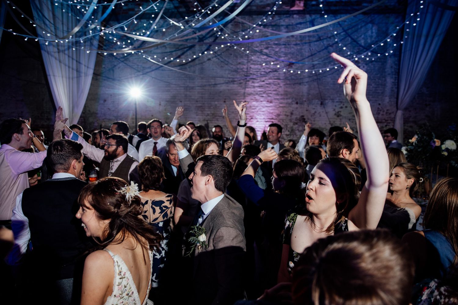 Crowded dance floor at wedding reception with blue lighting and white draping overhead.