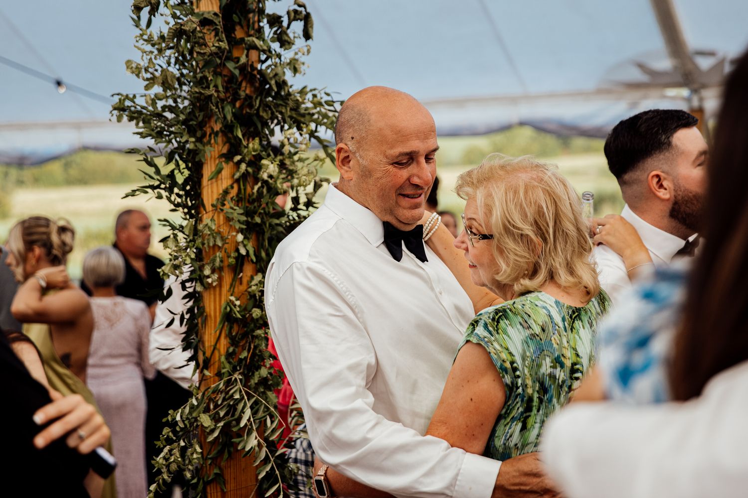 Wedding guests celebrate and dance together under an outdoor tent decorated with greenery.