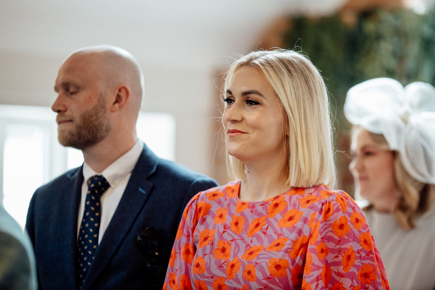 Wedding guests watching ceremony in bright indoor venue with decorative greenery.