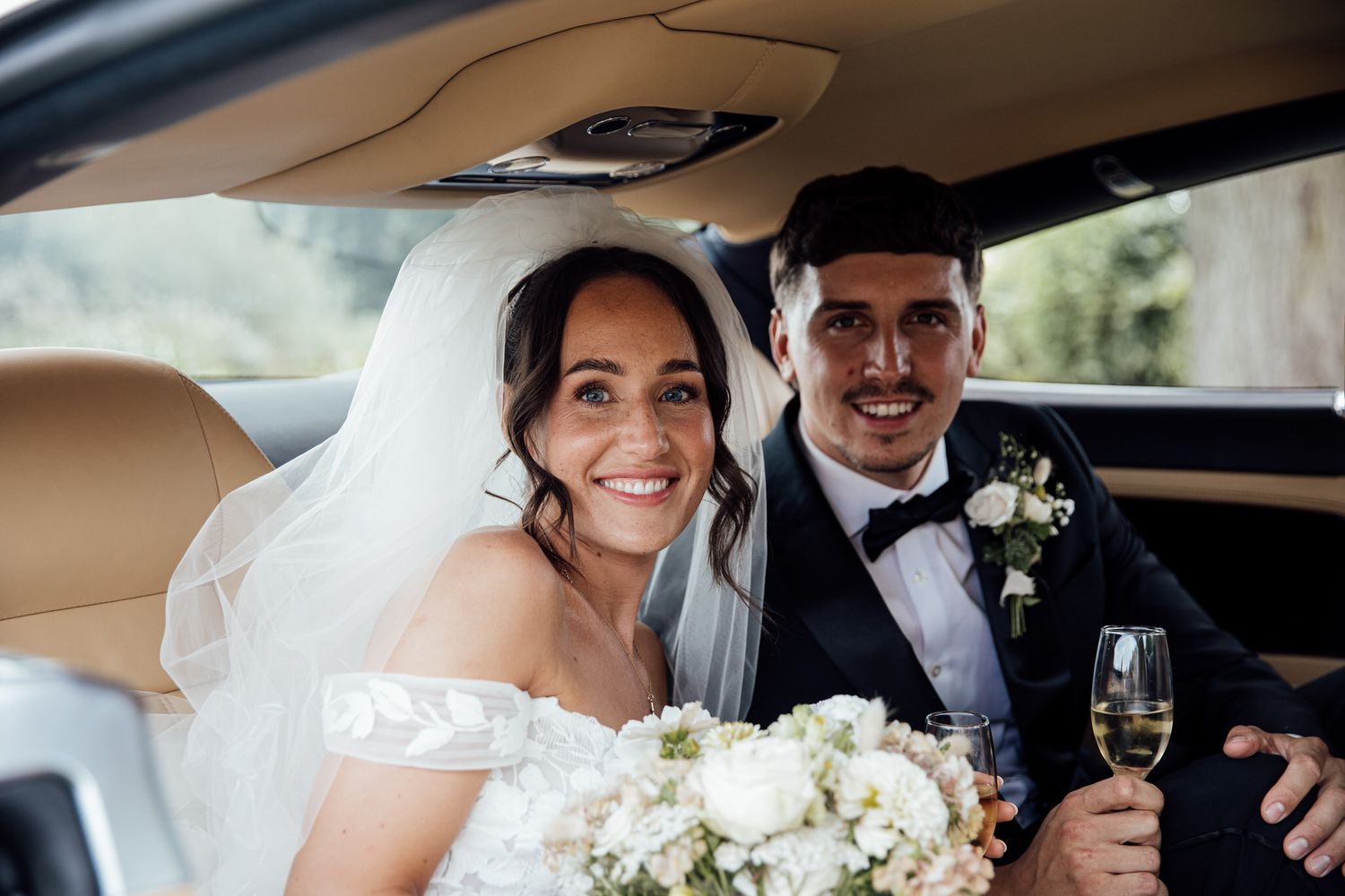 Newlyweds sharing champagne in the back of a luxury car with white floral bouquet.